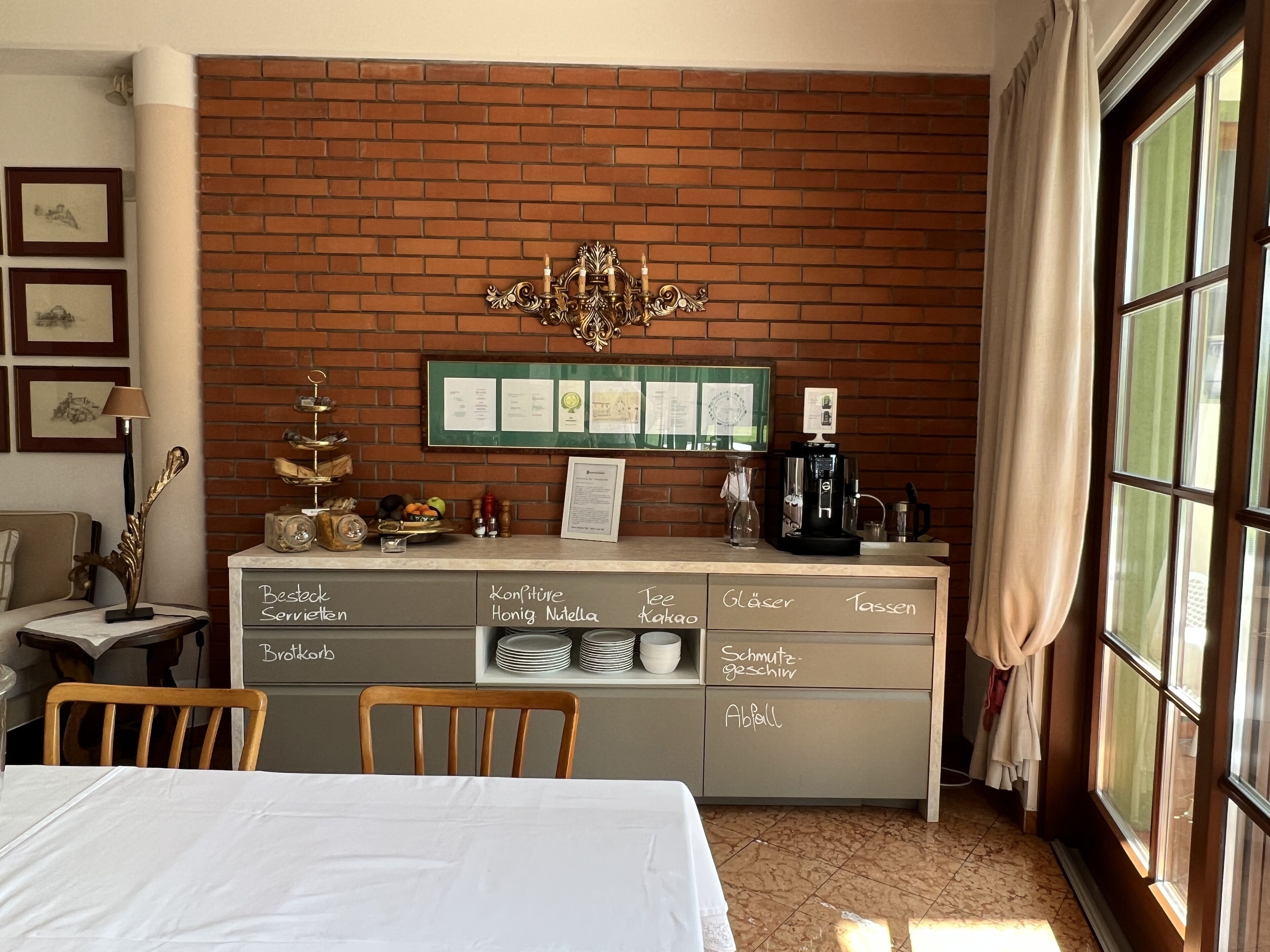 Ein Hotelzimmer mit Backsteinwand, einem Chandelier, einer Kaffeemaschine und einer Frühstückstheke mit beschrifteten Fächern.
