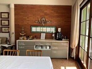 Une chambre d’hôtel avec un mur en briques, un lustre, une machine à café et un buffet pour le petit-déjeuner avec des compartiments étiquetés.
