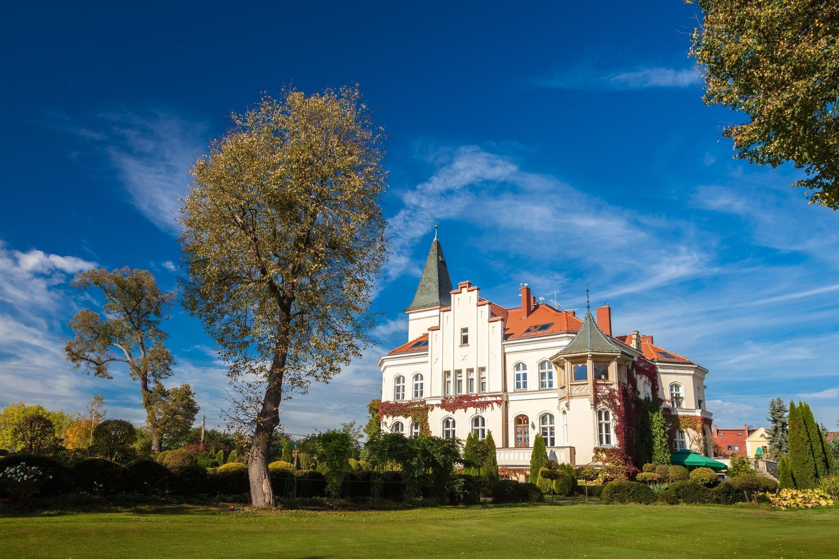 A large, historic building with a prominent tower, surrounded by well-maintained gardens and a clear blue sky.