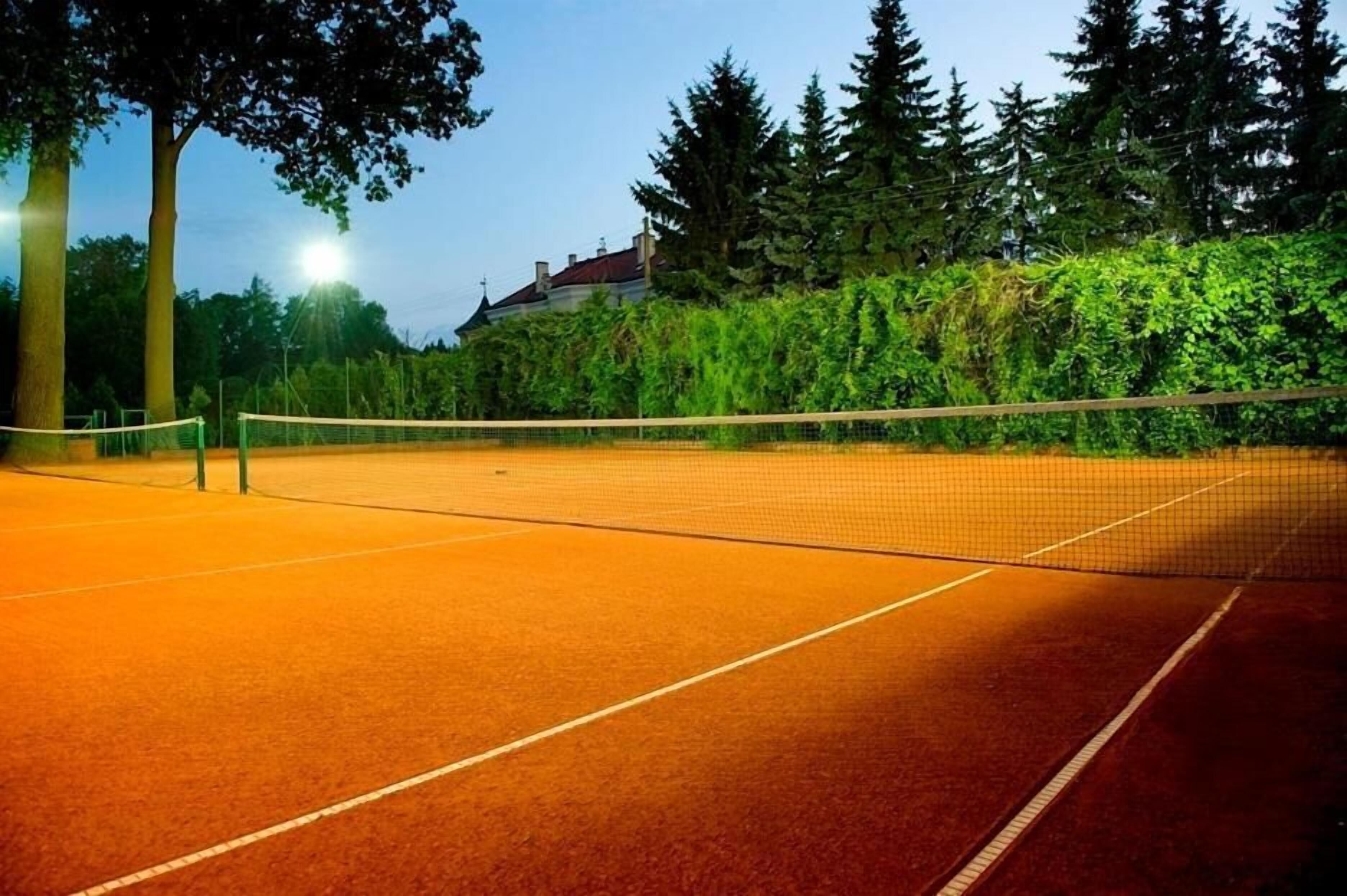 An outdoor tennis court with a net, surrounded by trees and a building in the background.