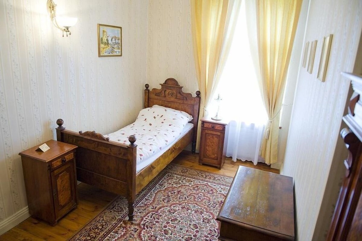 A bedroom with a wooden bed, a nightstand, a bedside table, a patterned rug, and a window with curtains.
