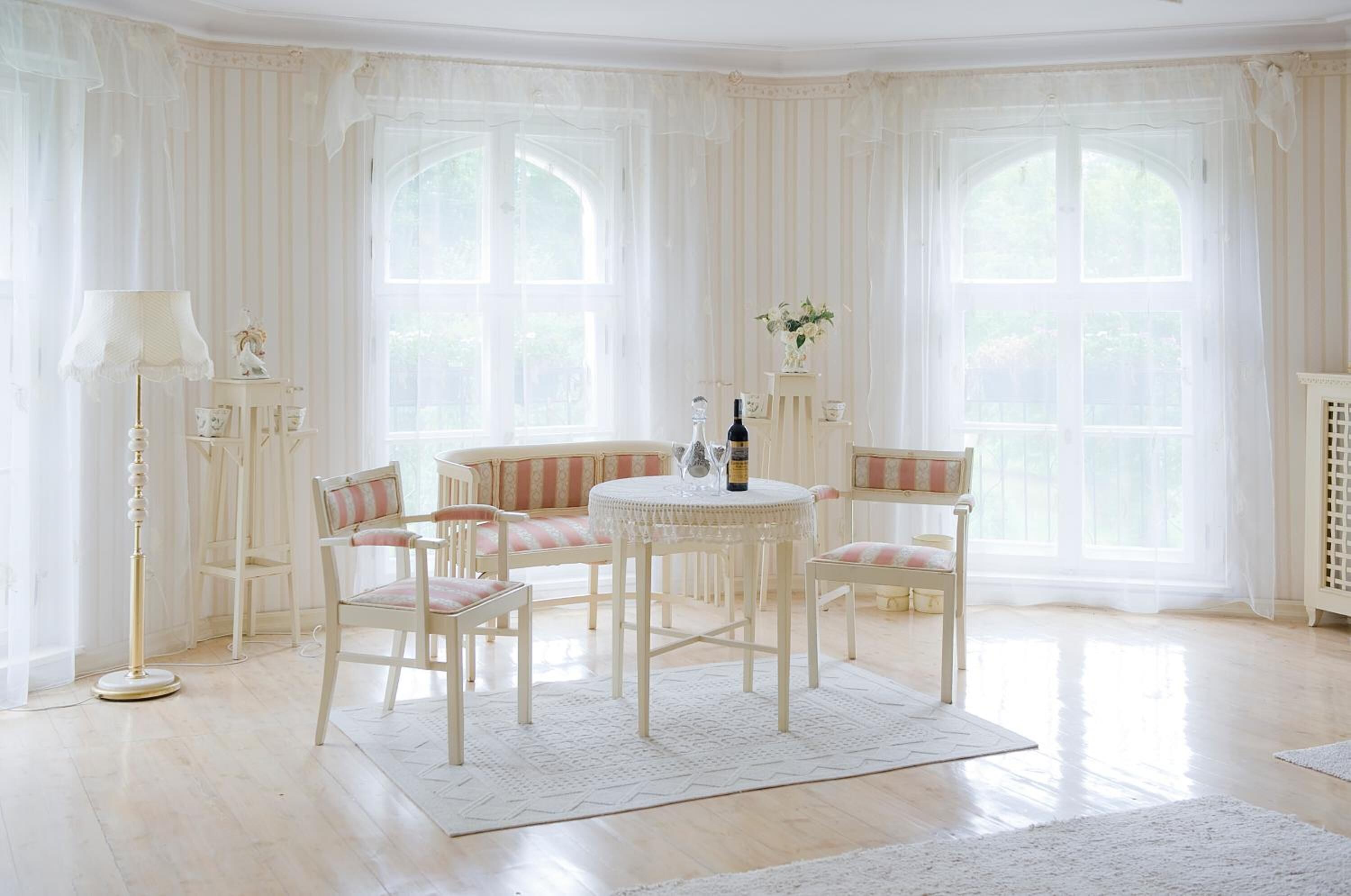 A room with a round table, four chairs, a floor lamp, and large windows with curtains.