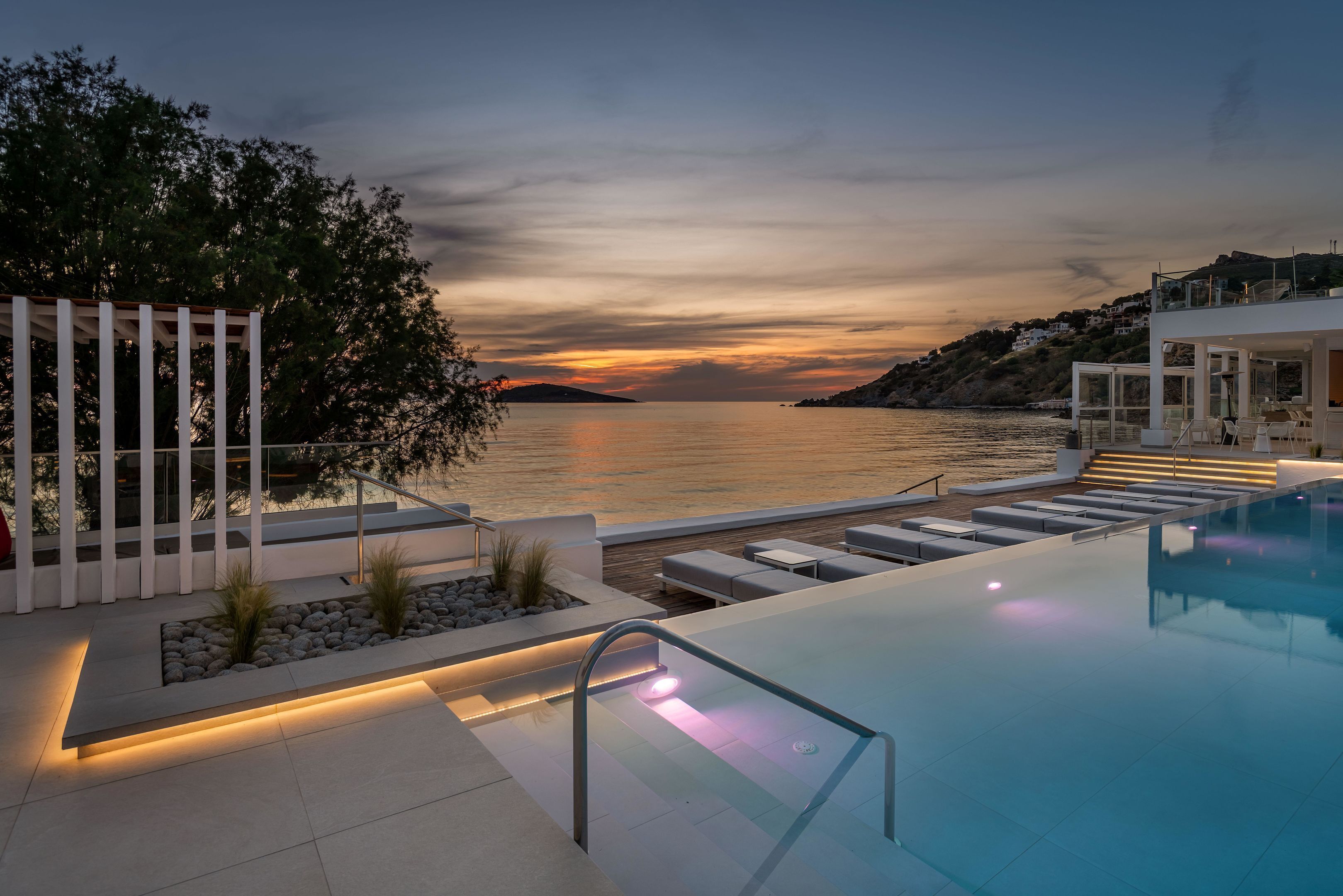 Un espace piscine extérieur moderne, doté d’une balustrade en verre, de chaises longues et offrant une vue sur la mer au coucher du soleil.