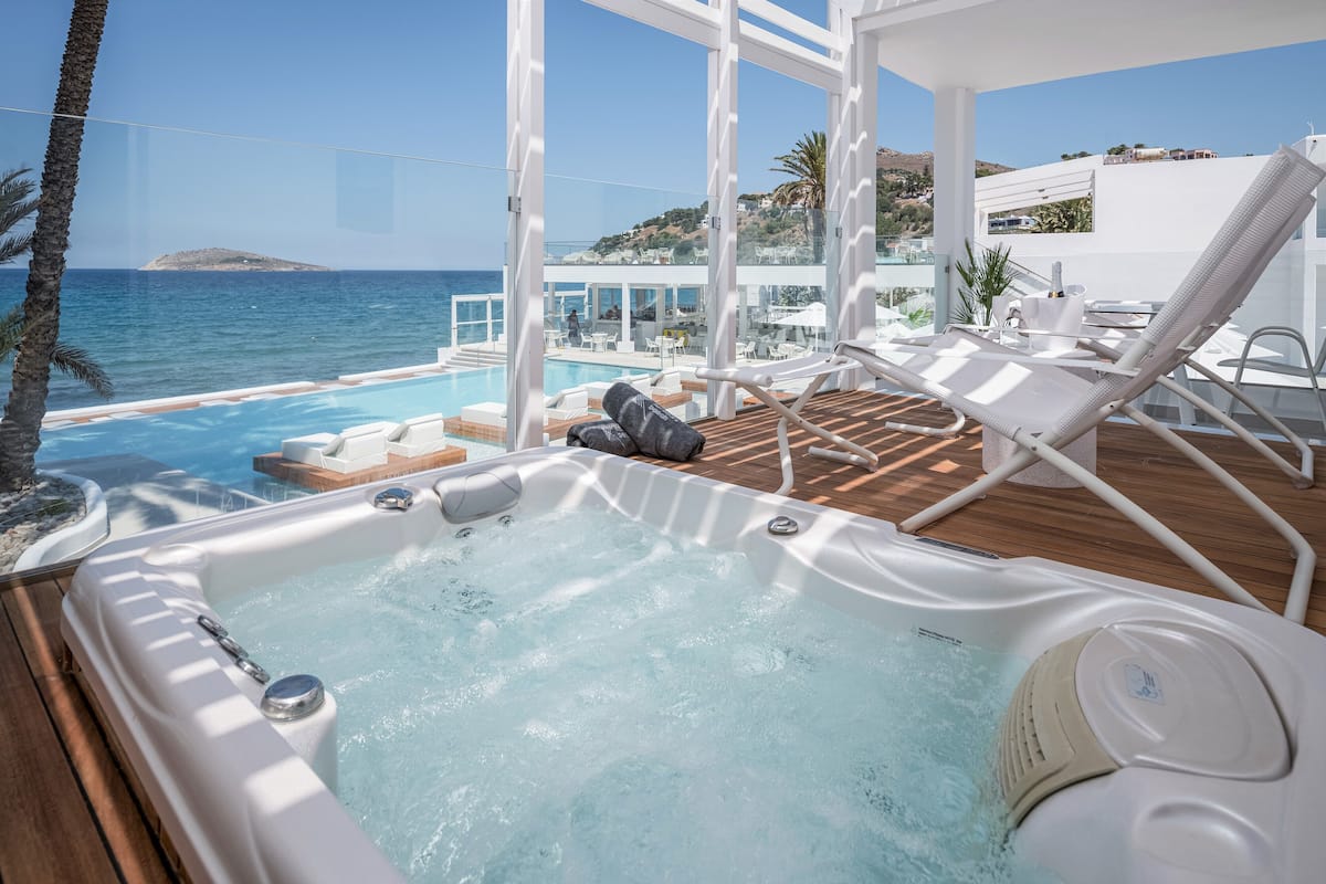 Ein Whirlpool auf einer Holzterrasse mit einem Pool und Liegestühlen, mit Blick auf das klare, blaue Meer und eine hügelige Landschaft.