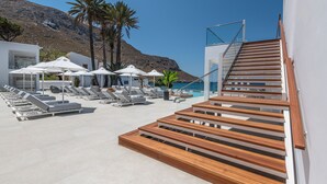 Un área de piscina en la azotea con tumbonas y sombrillas, una escalera de madera y vistas al mar y a las montañas.