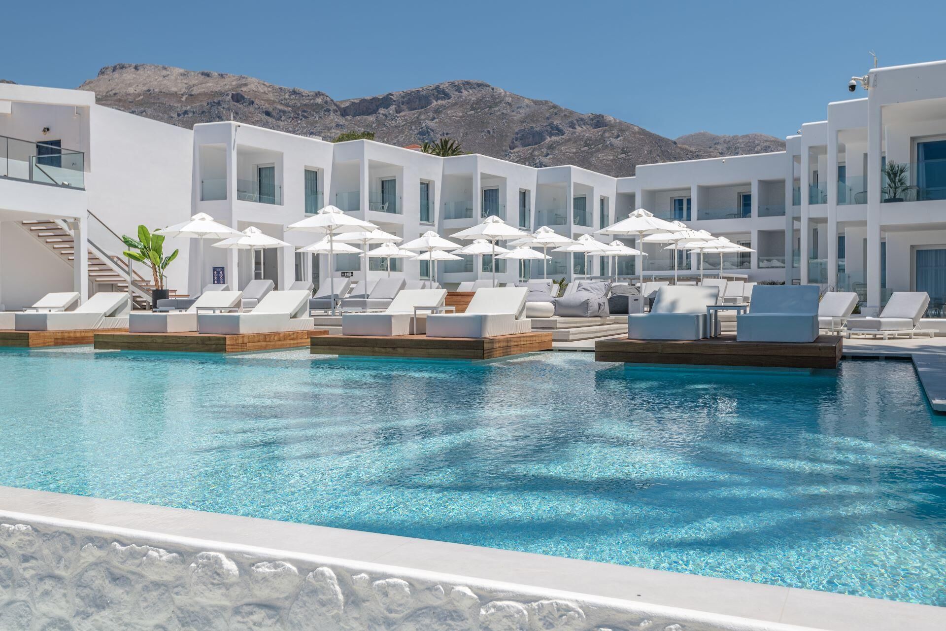 A pool area with sun loungers and umbrellas, surrounded by white buildings with large windows, set against a mountainous backdrop.