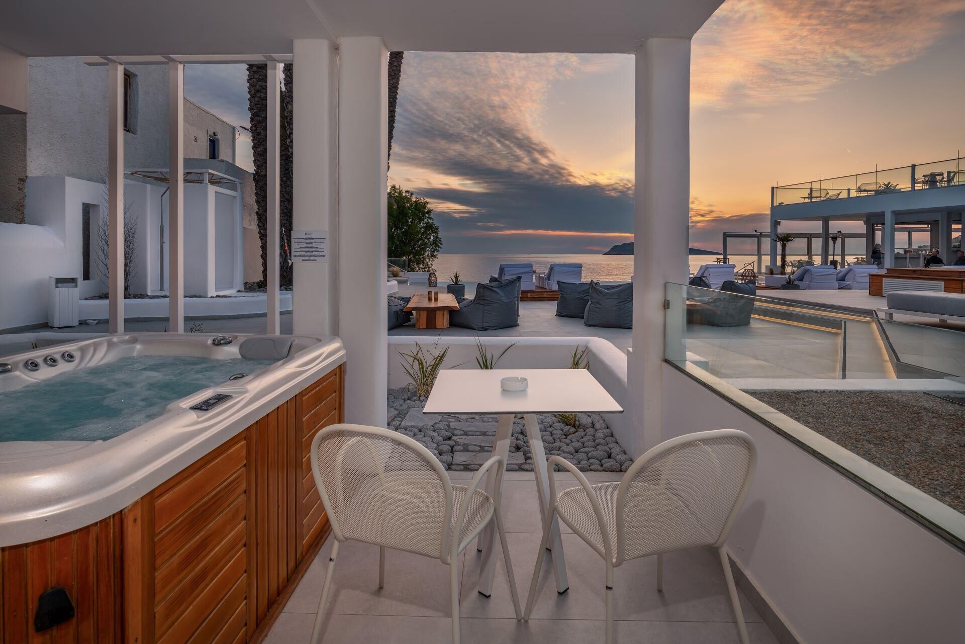 A modern outdoor seating area with a hot tub, white chairs, and a table, overlooking a body of water during sunset.