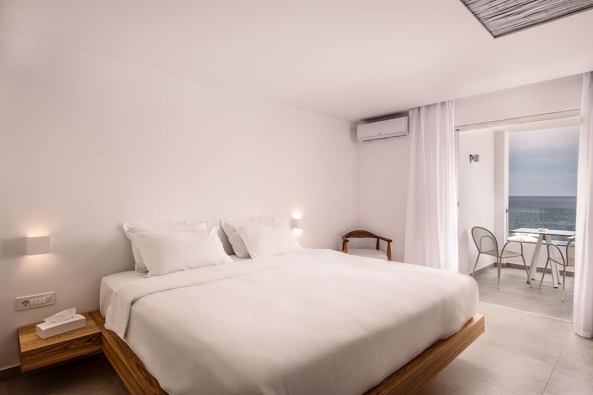 Ein modernes Hotelzimmer mit einem großen Bett, einem Nachttisch und Blick aufs Meer durch eine geöffnete Tür.