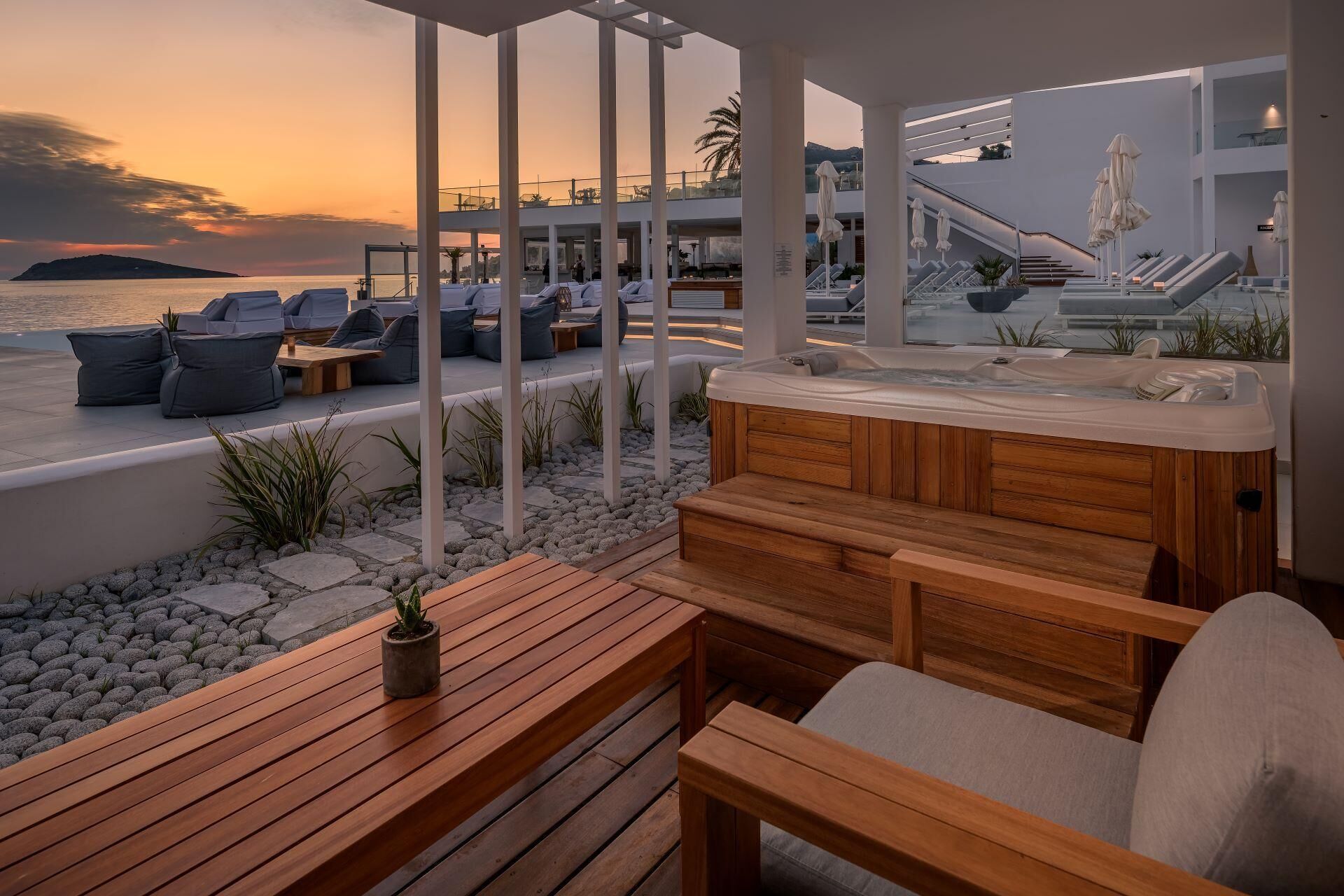 Un área de estar exterior moderna con un jacuzzi, asientos de madera y vistas al mar al atardecer.