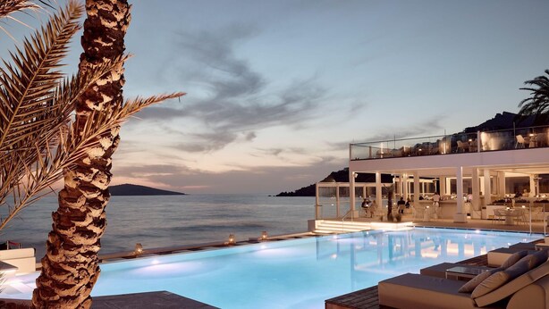 Una piscina infinita moderna con vista despejada al mar, un área de descanso con asientos y un comedor envuelto en vidrio.