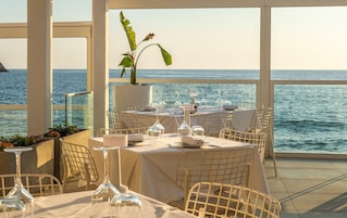 Mesa lista para comer con vista al mar, sillas blancas y una planta en maceta.