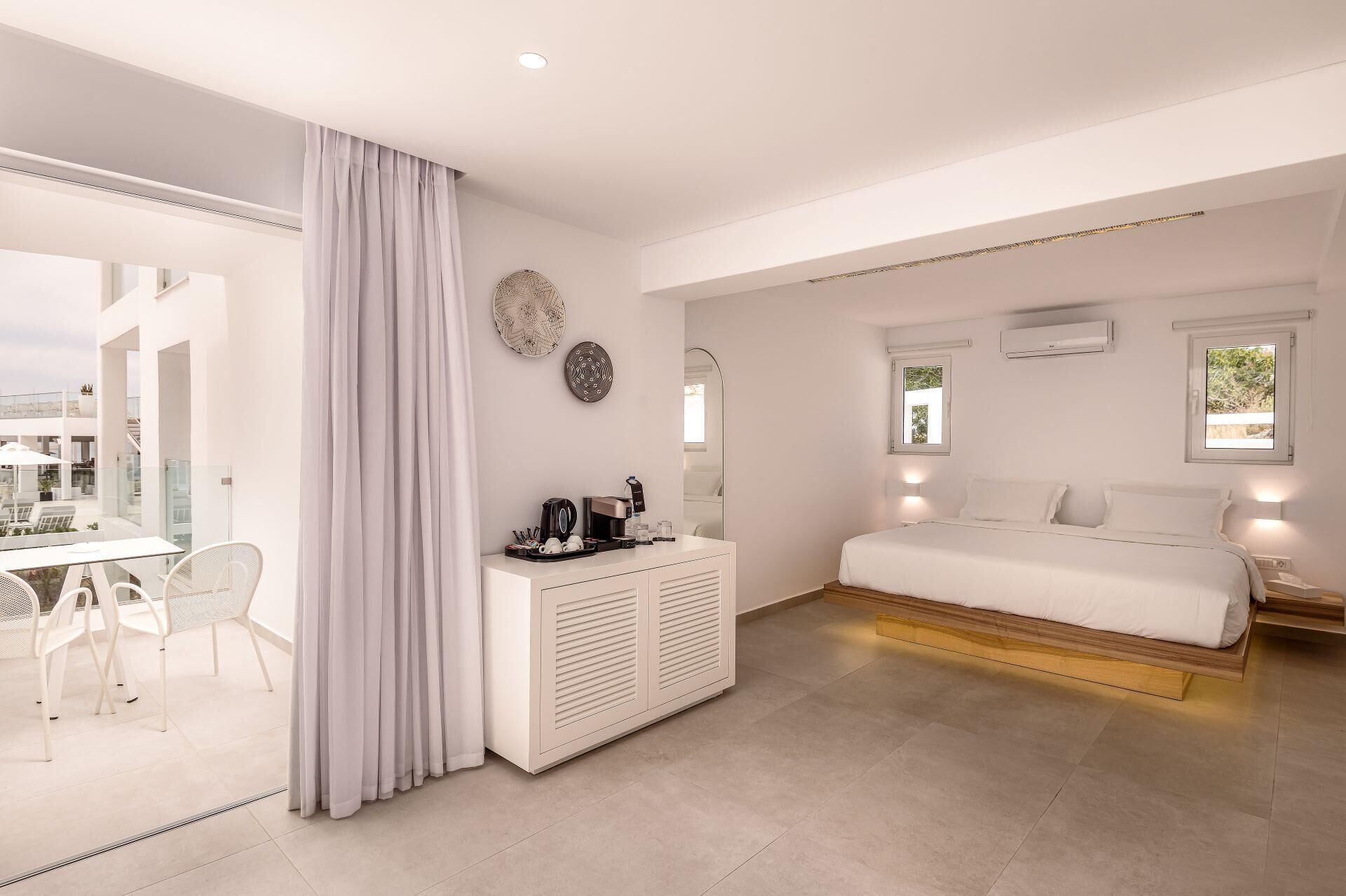 Una habitación de hotel moderna con una cama blanca, un escritorio pequeño blanco y un área de comedor con sillas blancas.