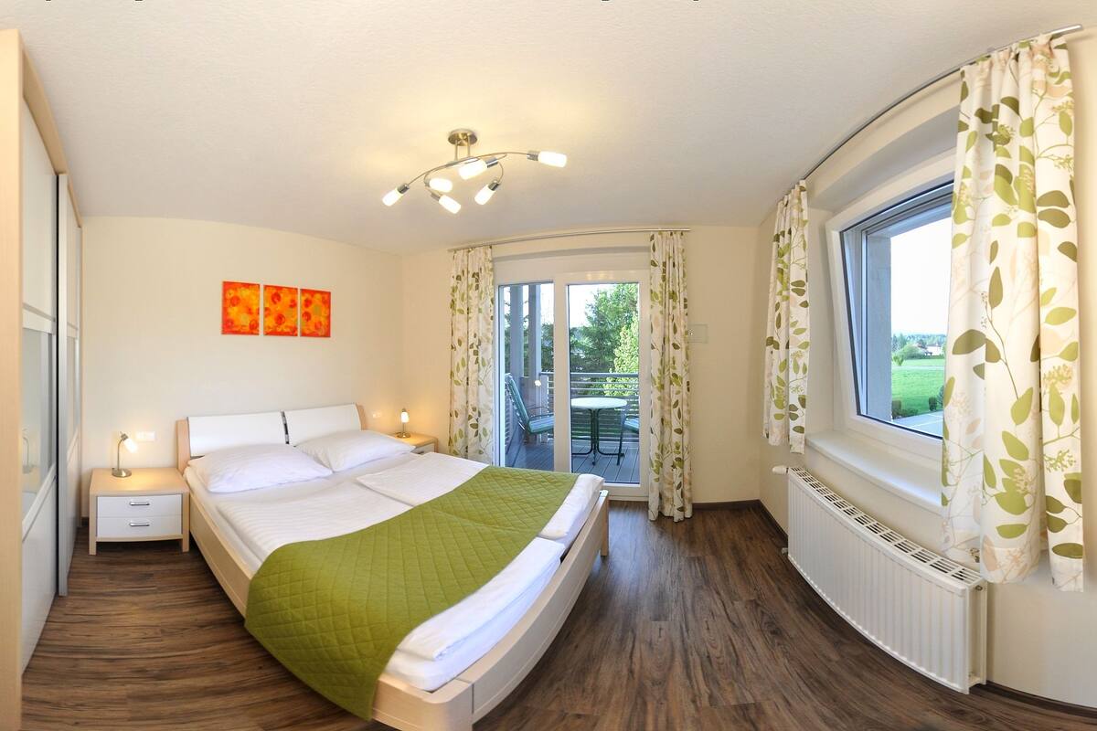 Ein Schlafzimmer mit Bett, Nachttischen, einem Fenster mit Vorhängen, einem Balkon und einer Deckenleuchte.