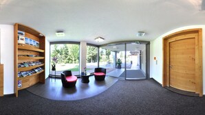 A reception area with a wooden reception desk, a seating area with two chairs, a large glass door leading outside, and a wooden door on the right. - Karglhof - Stammhaus (Finkenstein am Faaker See)