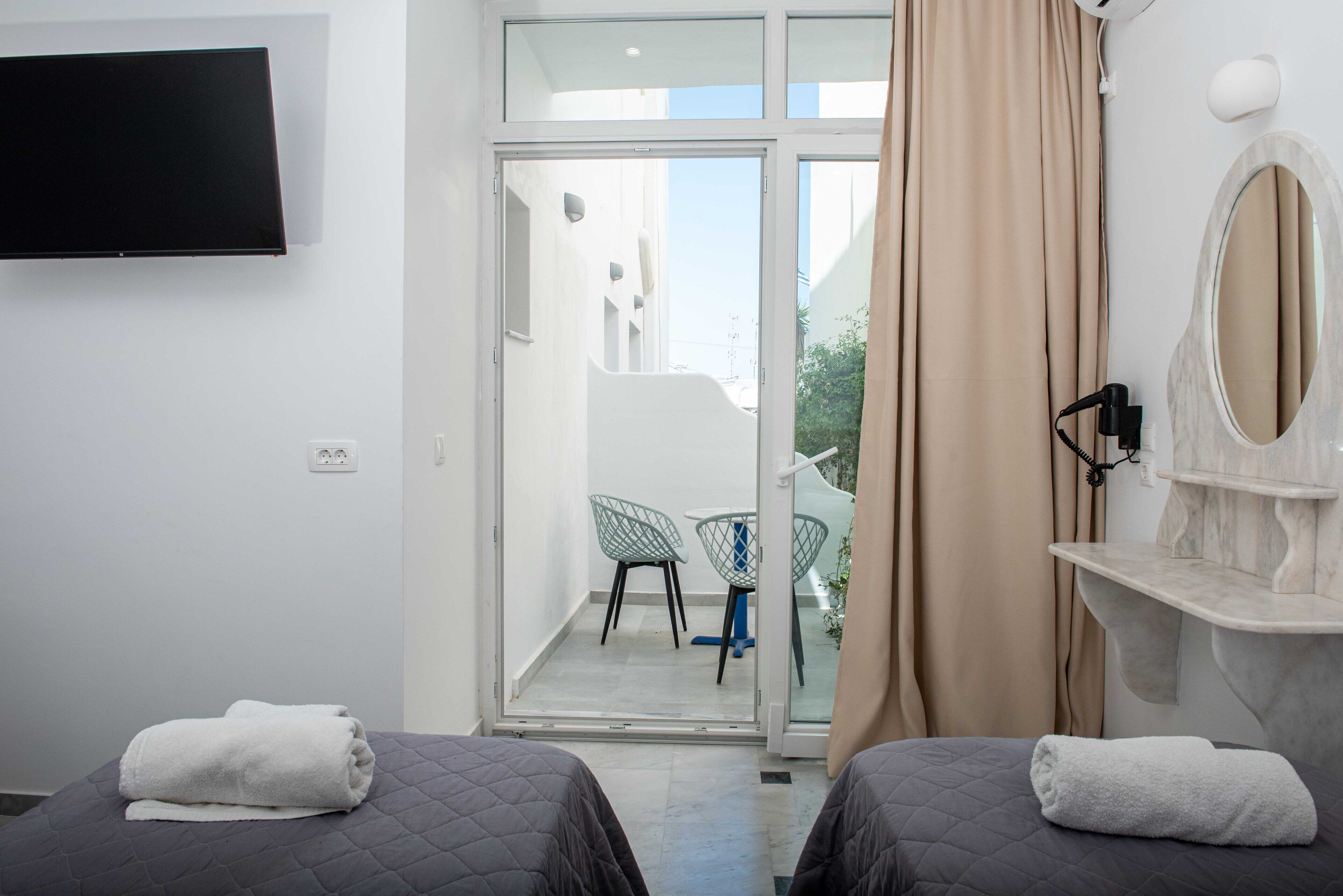 A hotel room with two beds, a TV, a balcony with chairs, and a bathroom with a mirror and towel rack.
