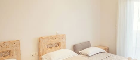 Camera d'albergo con tre letti, ognuno dotato di testiera in legno, biancheria da letto bianca e asciugamani arrotolati. Un comodino è posizionato tra i letti, e una finestra è ornata da tende.