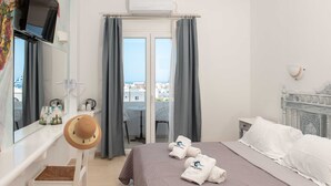 Habitación de hotel con cama, escritorio con silla, televisión, ventana con cortinas y vistas a edificios.