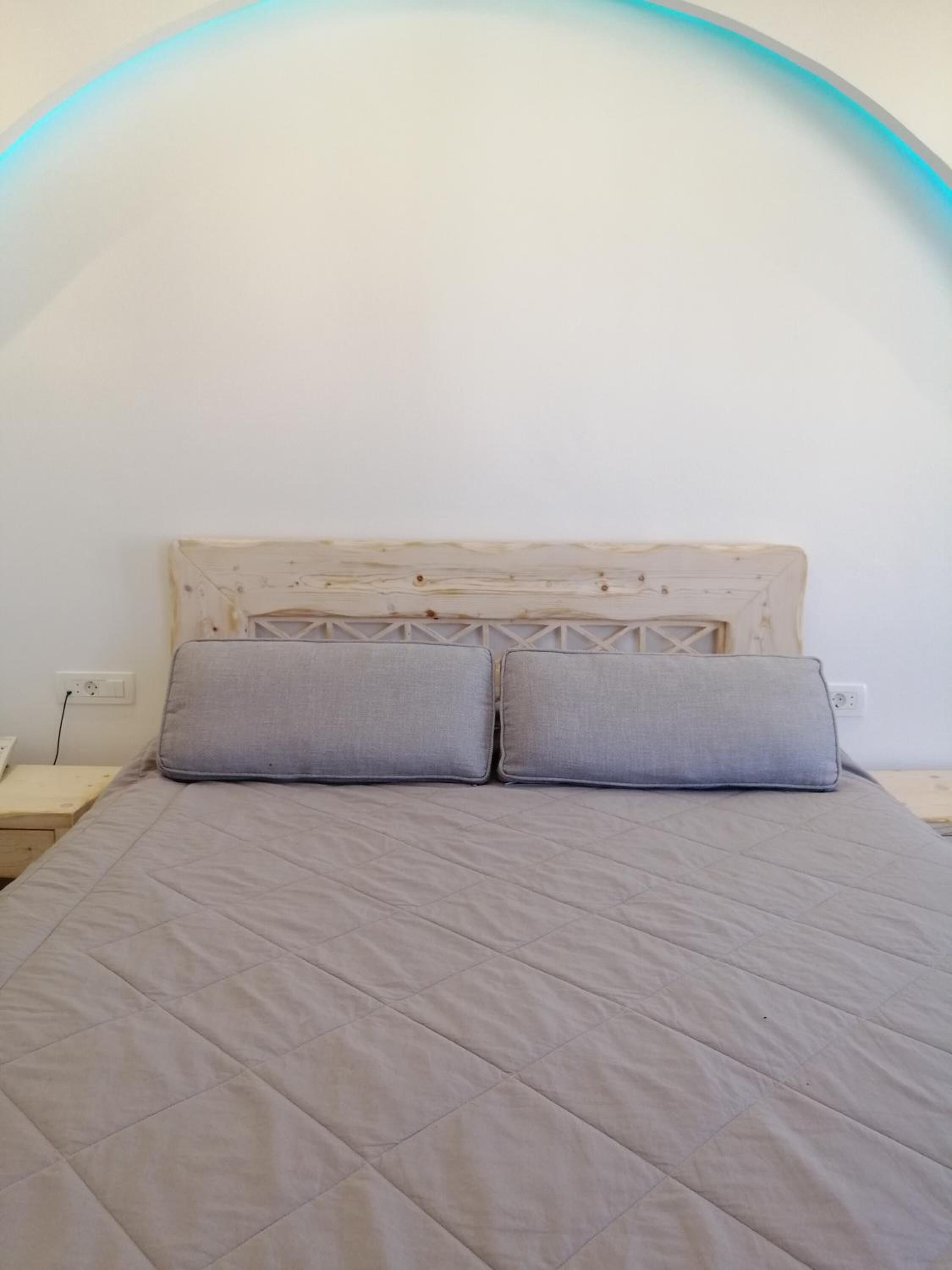 Een bed met een gewatteerde grijze sprei en twee grijze kussens, een houten hoofdbord, en een witte muur met een blauwe lichtboog boven het bed.