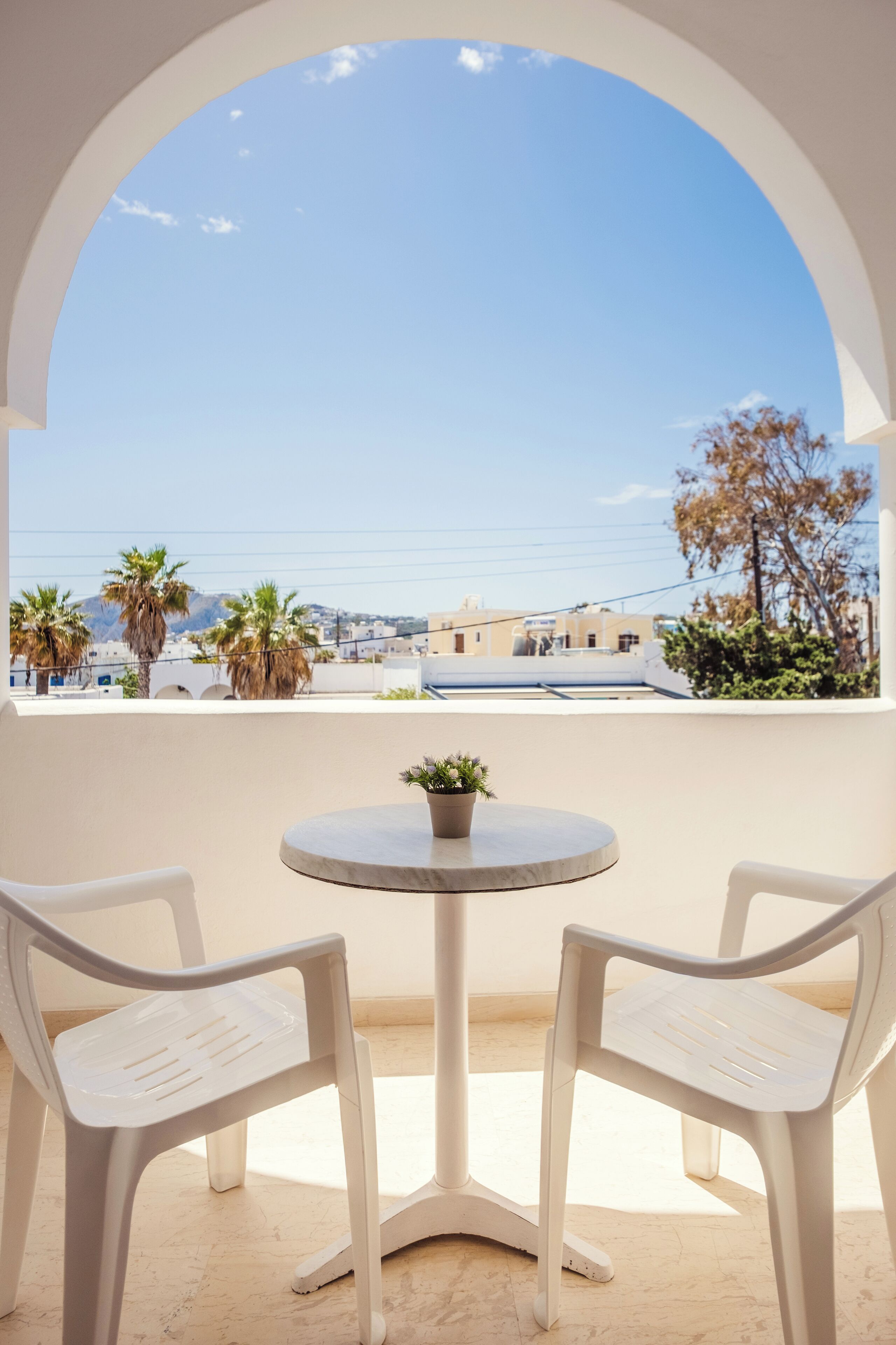A balcony with a round table and two white chairs, overlooking a cityscape with buildings and palm trees.
