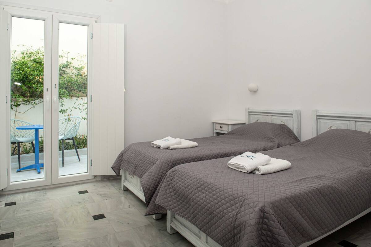 A hotel room with two beds, a small table, and a chair by a sliding glass door leading to an outdoor area.