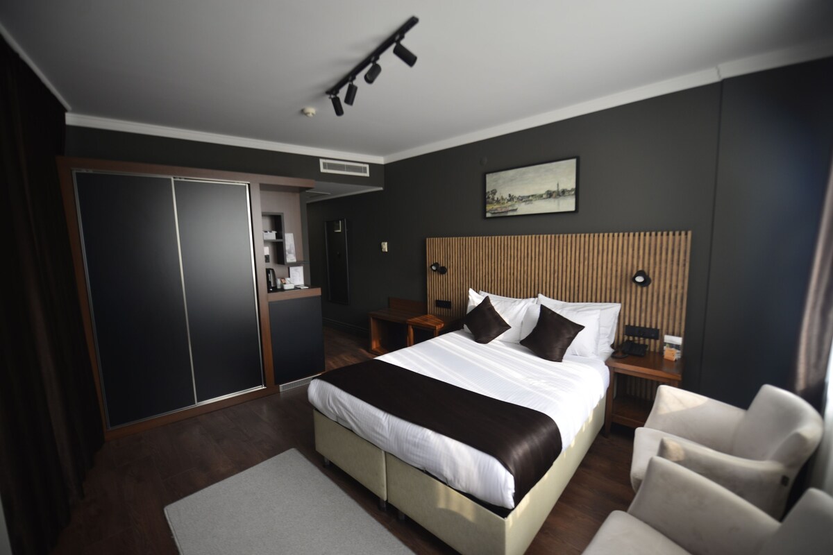 A modern hotel room with a large bed, a desk, a chair, a wardrobe, and a painting on the wall.