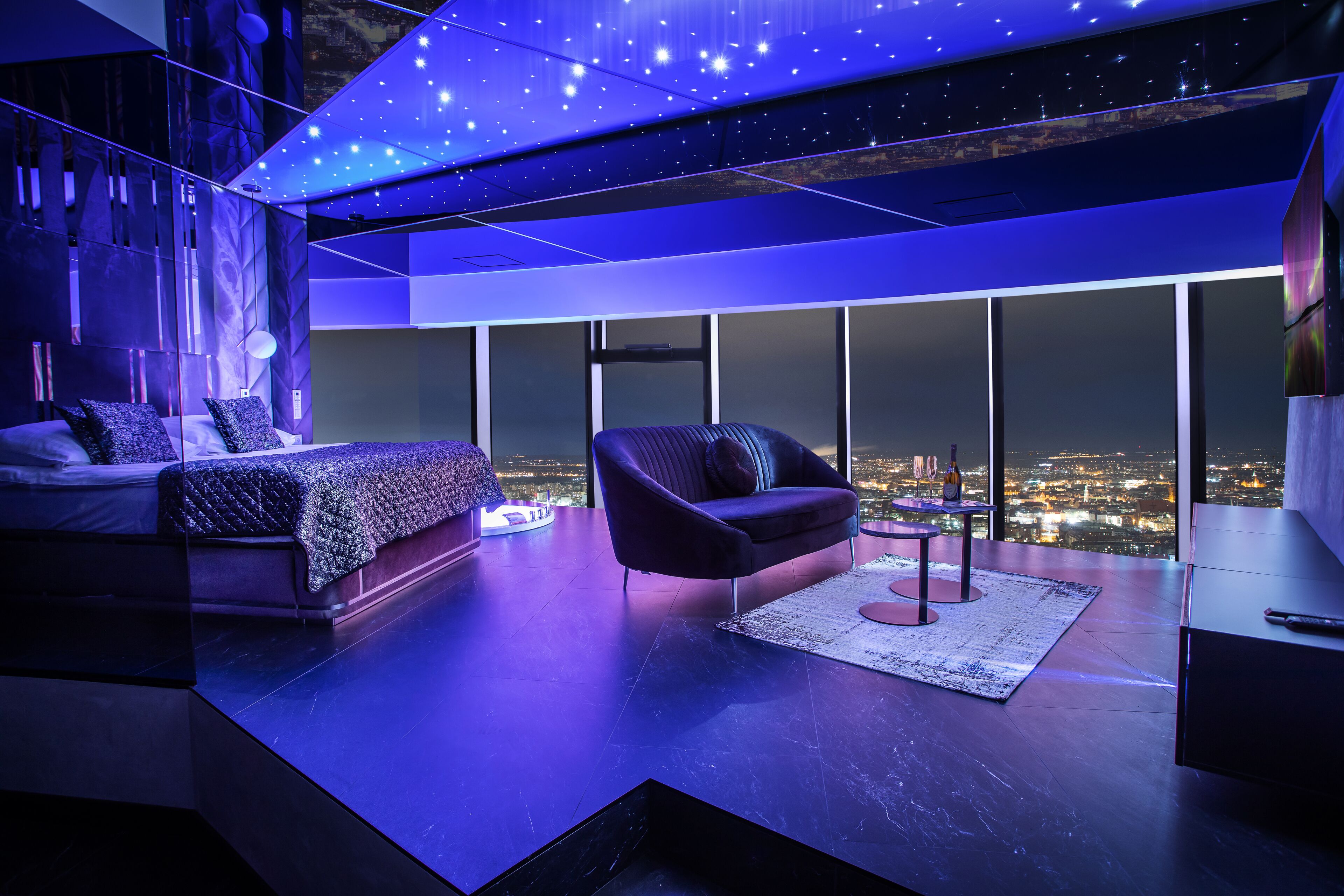 A modern bedroom with a city view, a large bed, a sofa, and a desk.