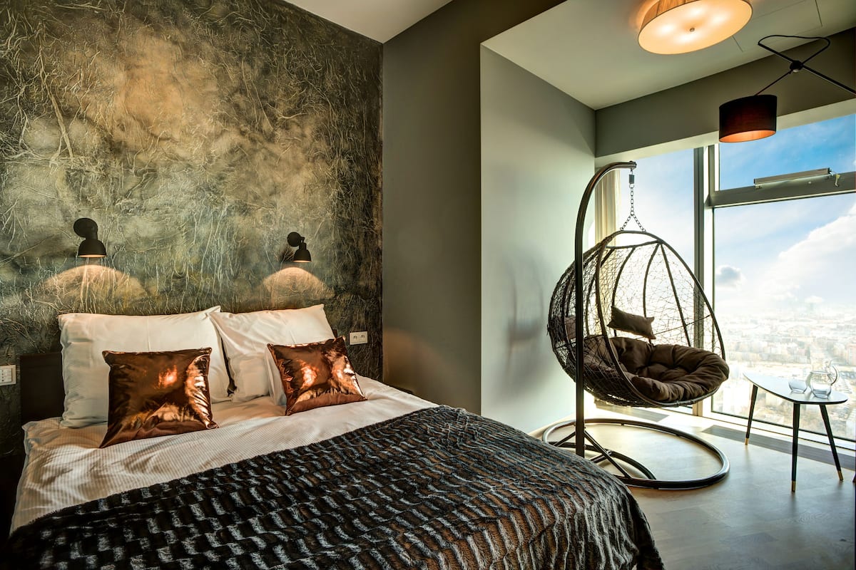 Ein modernes Schlafzimmer mit einem großen Bett, einem Hängesessel und Blick auf die Stadt durch das Fenster.