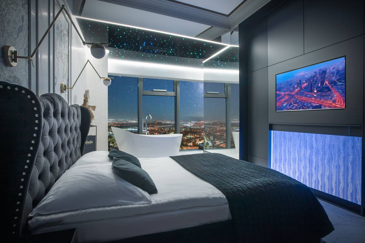 Ein modernes Schlafzimmer mit einem großen Bett, Blick auf die nächtliche Stadt und einem großen Bildschirm an der Wand.