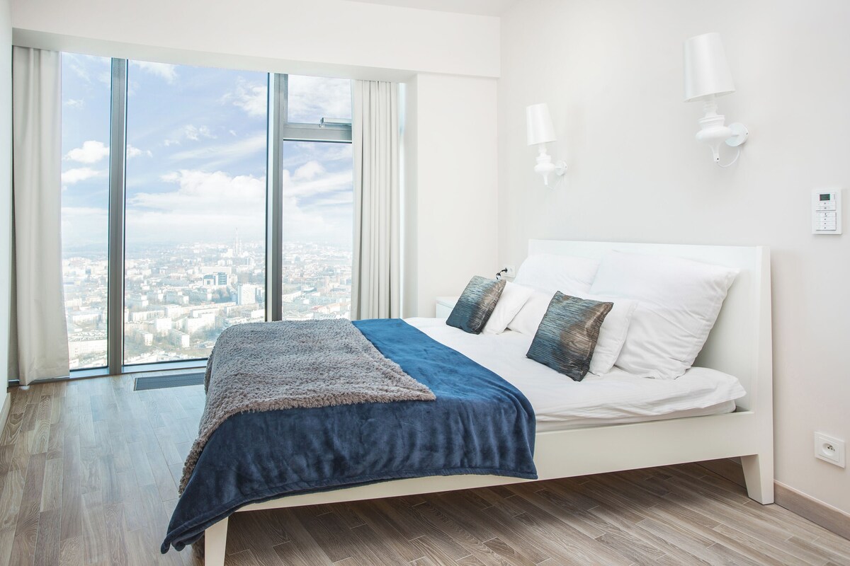 A modern bedroom with a large bed, a city view, and a white wall with a light fixture.