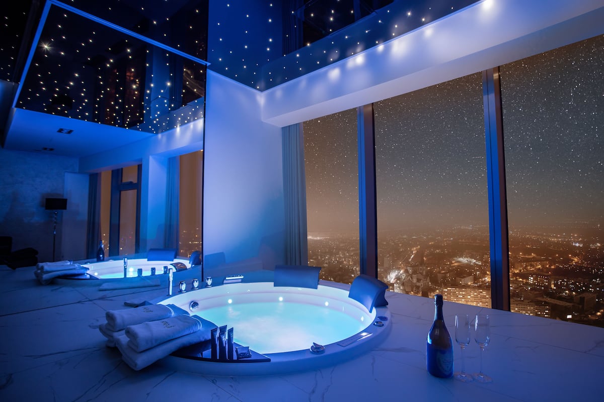 Ein luxuriöses Zimmer mit Whirlpool, einer Champagnerflasche und Gläsern auf einem Tisch, mit Blick auf die Stadt durch große Fenster.