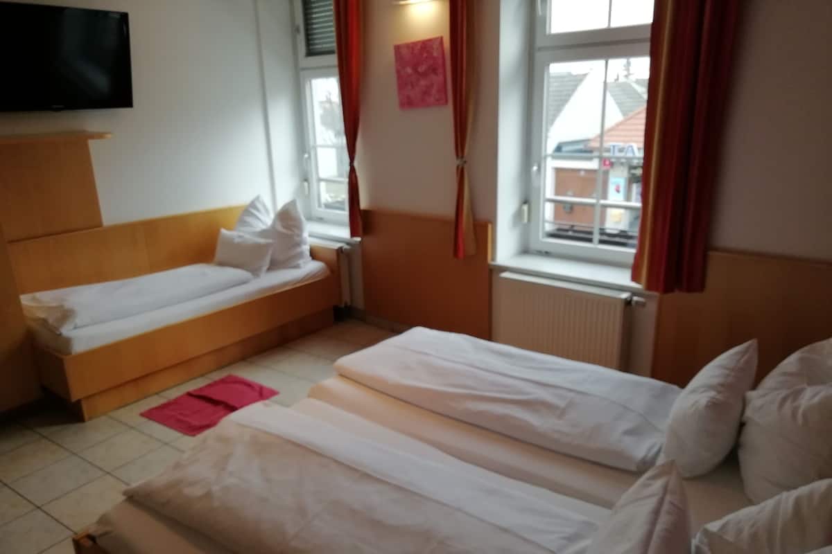 A hotel room with two single beds, a wooden headboard, a flat-screen TV, a window with curtains, and a radiator.