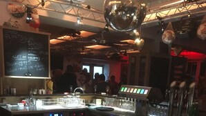 Een barruimte met een toonbank, krukken en een discobal. Er is een krijtbordmenu en een groep mensen op de achtergrond.