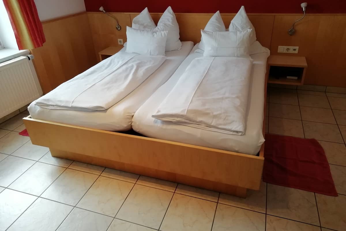 A double bed with white linens, a wooden headboard, a nightstand with a lamp, and a red curtain.