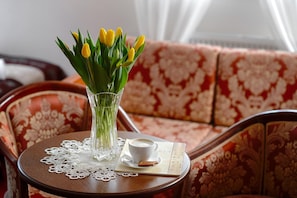 A clear glass vase with yellow tulips on a small round table with a lace doily, a white cup, and a biscuit on a napkin beside a patterned sofa.