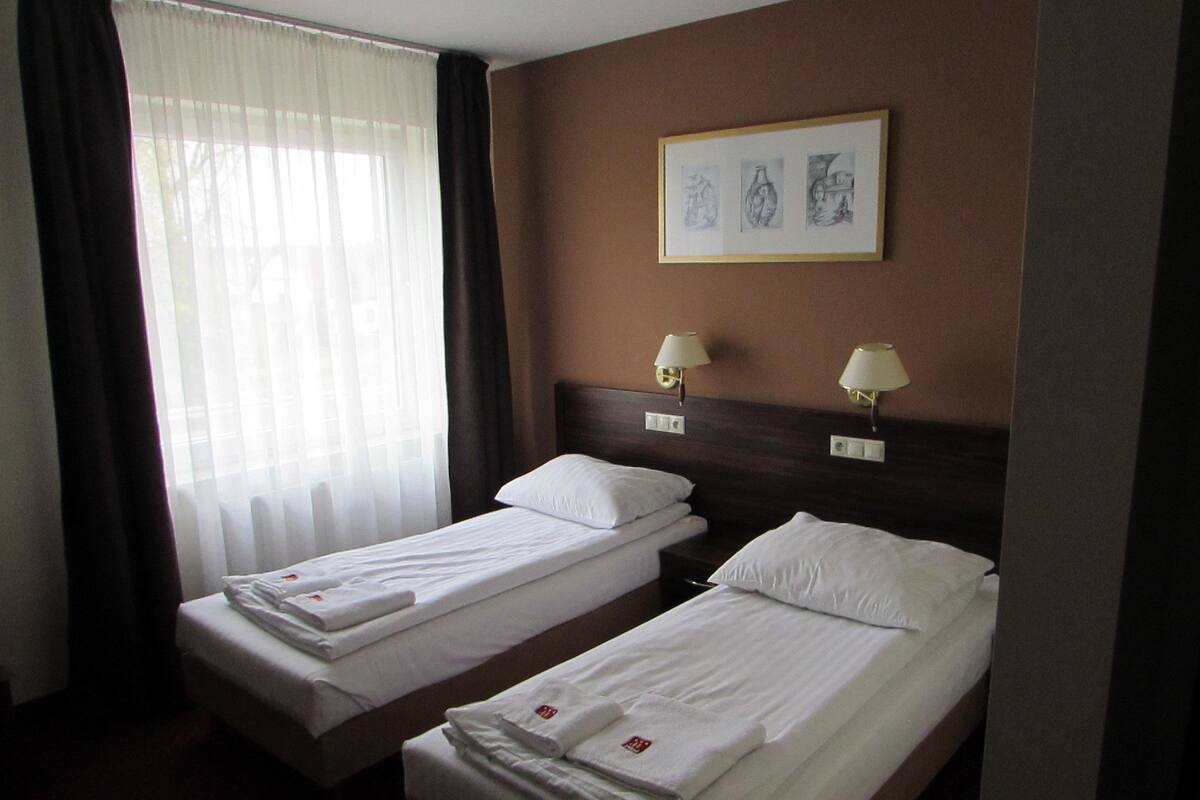 A hotel room with two single beds, a window with sheer curtains, and a framed artwork on the wall.
