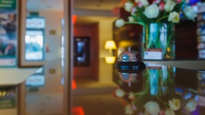 En hotellobby med en välpolerad receptionsdisk, en vas med blommor och en inramad tavla på väggen.