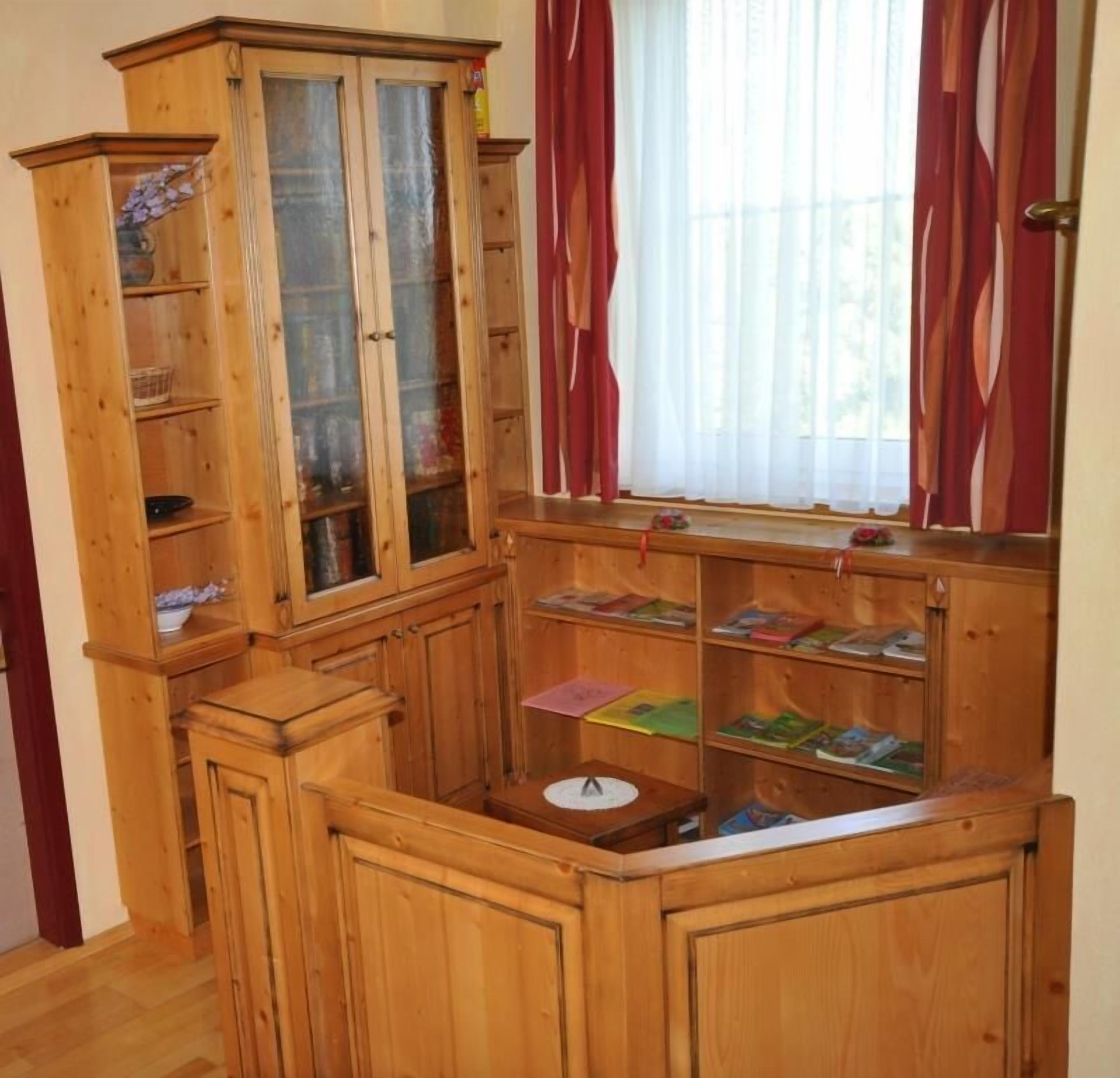 Un mueble de madera con varios estantes y cajones, una pequeña mesa con mantel blanco y una ventana con cortinas rojas.