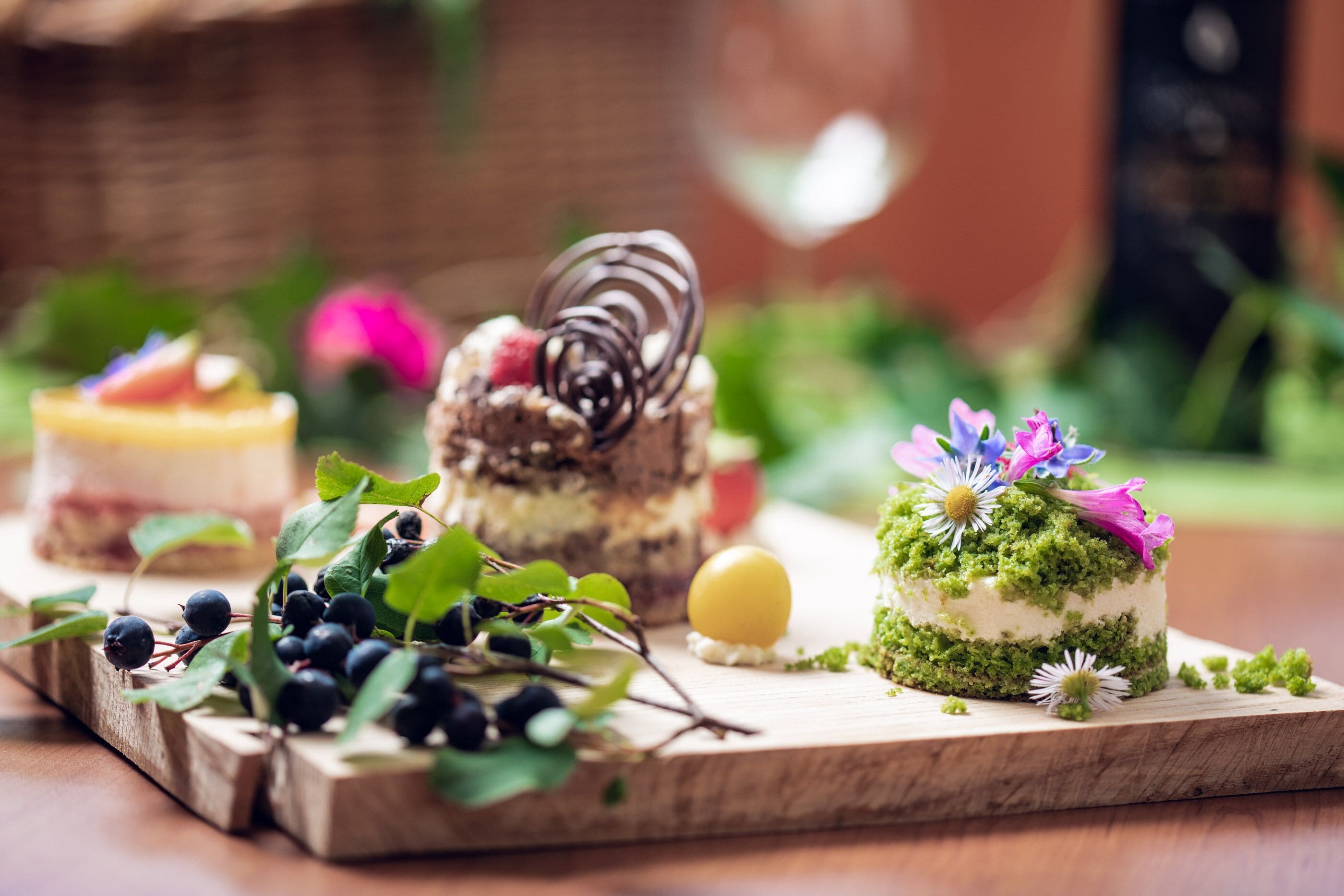 A wooden board with various desserts, including a cake with a spiral chocolate decoration and a cake with green frosting and flowers.