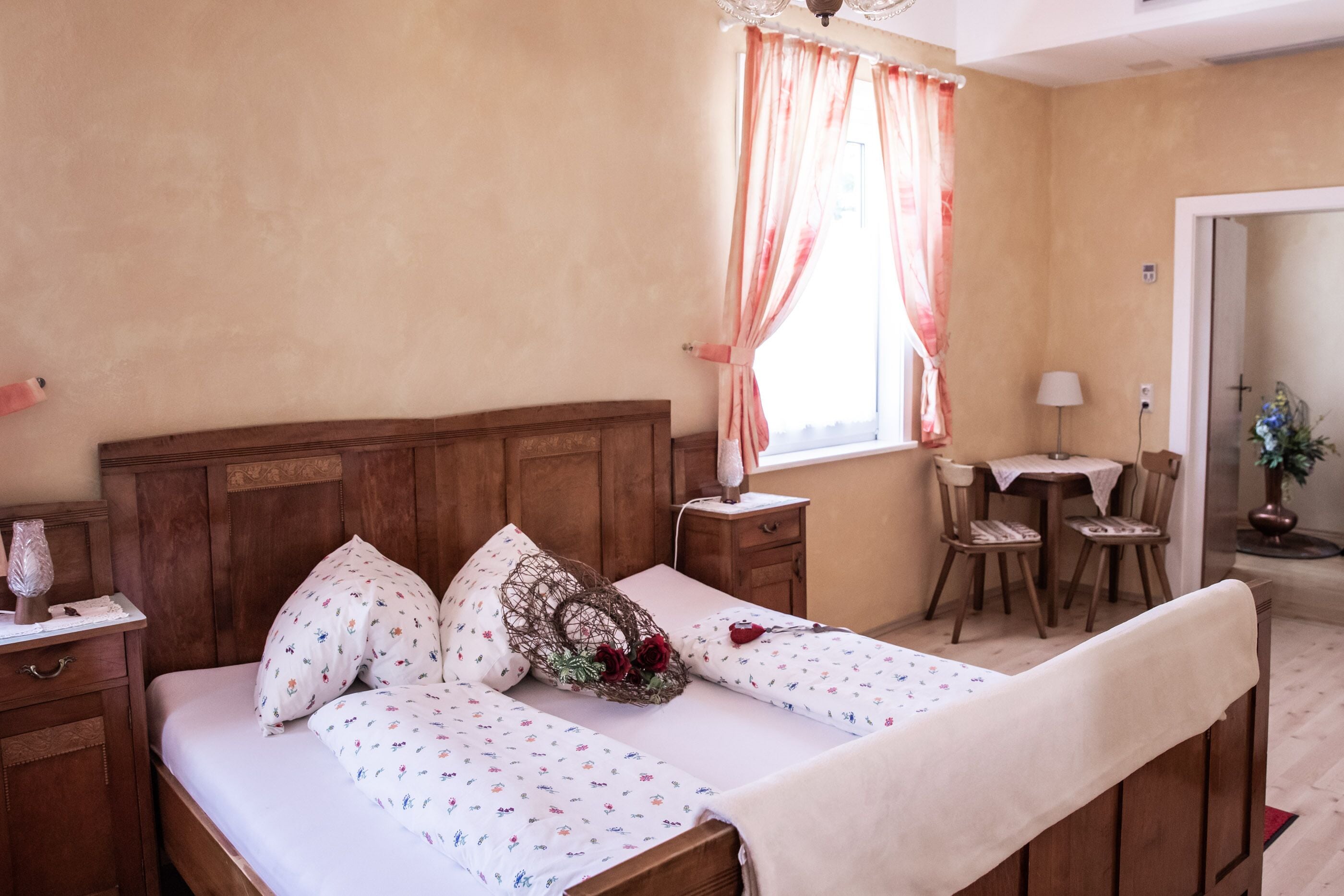 A bedroom with a wooden bed, floral bedding, a bedside table with a lamp, and a window with curtains.