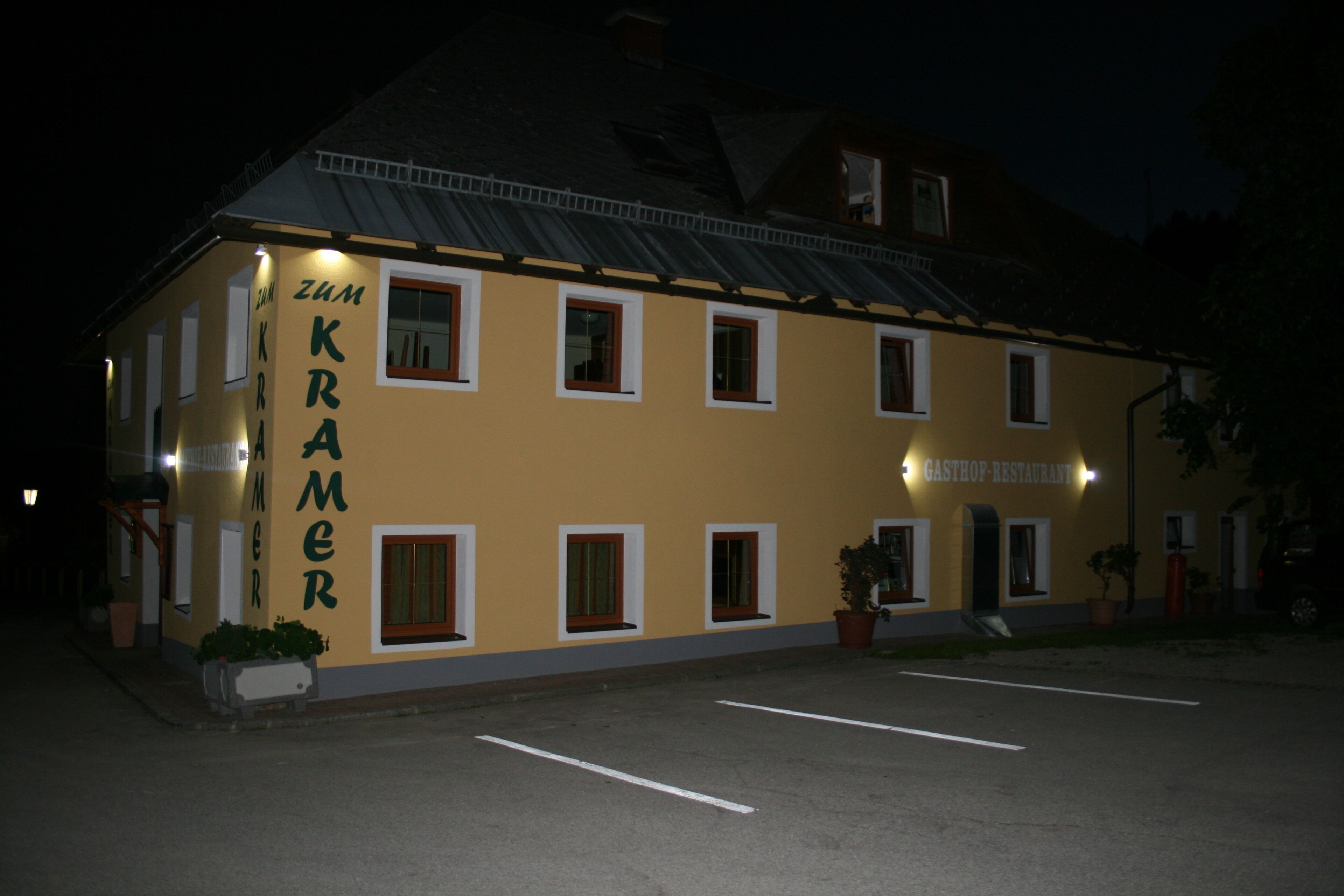 A building with the sign "Zum Kramer" and "Gasthof Restaurant" indicating it is a restaurant. There are multiple windows and a door, and the building is lit at night.