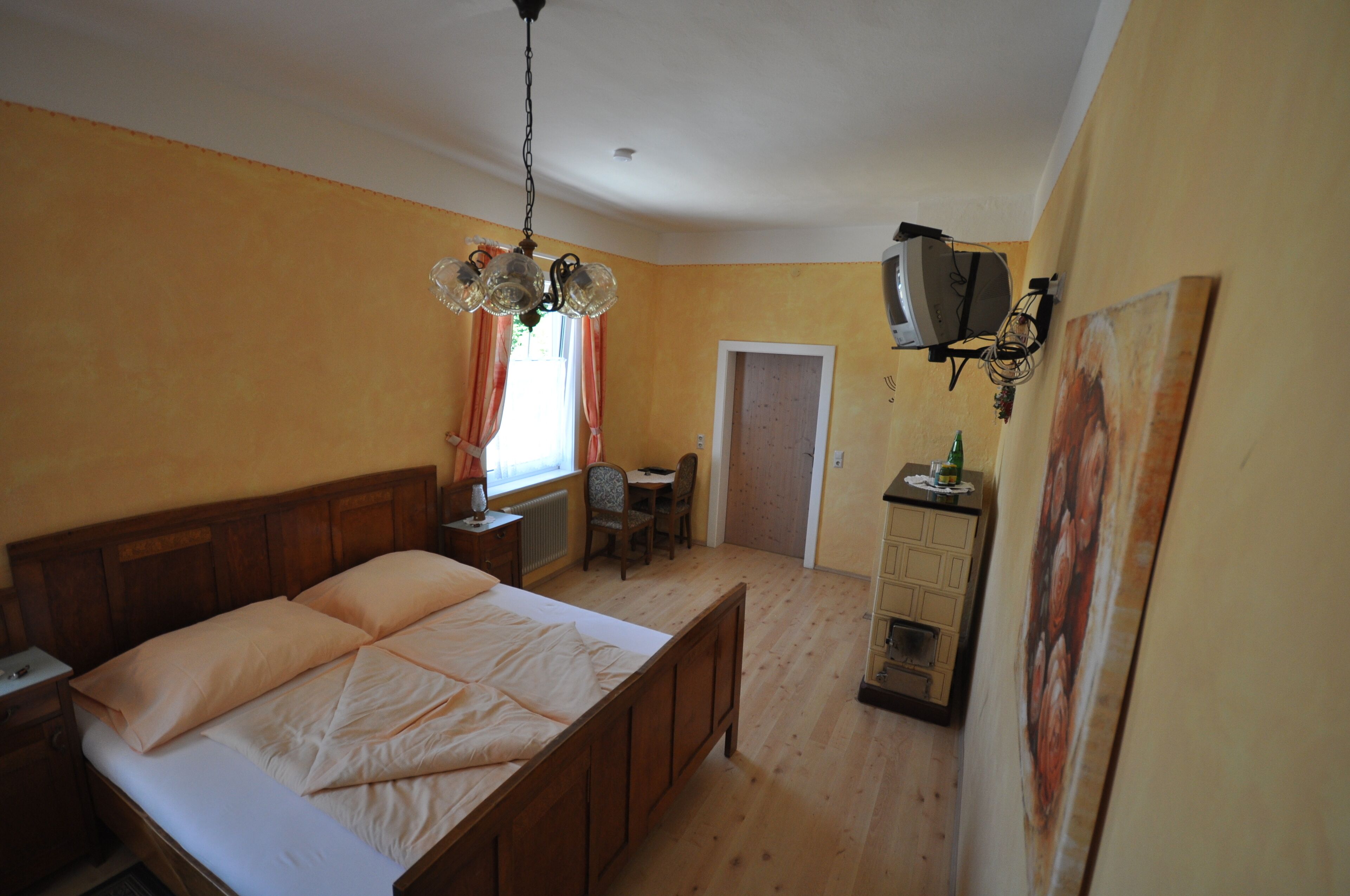 Ein Schlafzimmer mit einem hölzernen Bett, einem Schreibtisch und einem Stuhl. Es gibt ein Fenster mit Vorhängen, eine Heizung und ein an der Wand befestigtes Fernsehgerät.