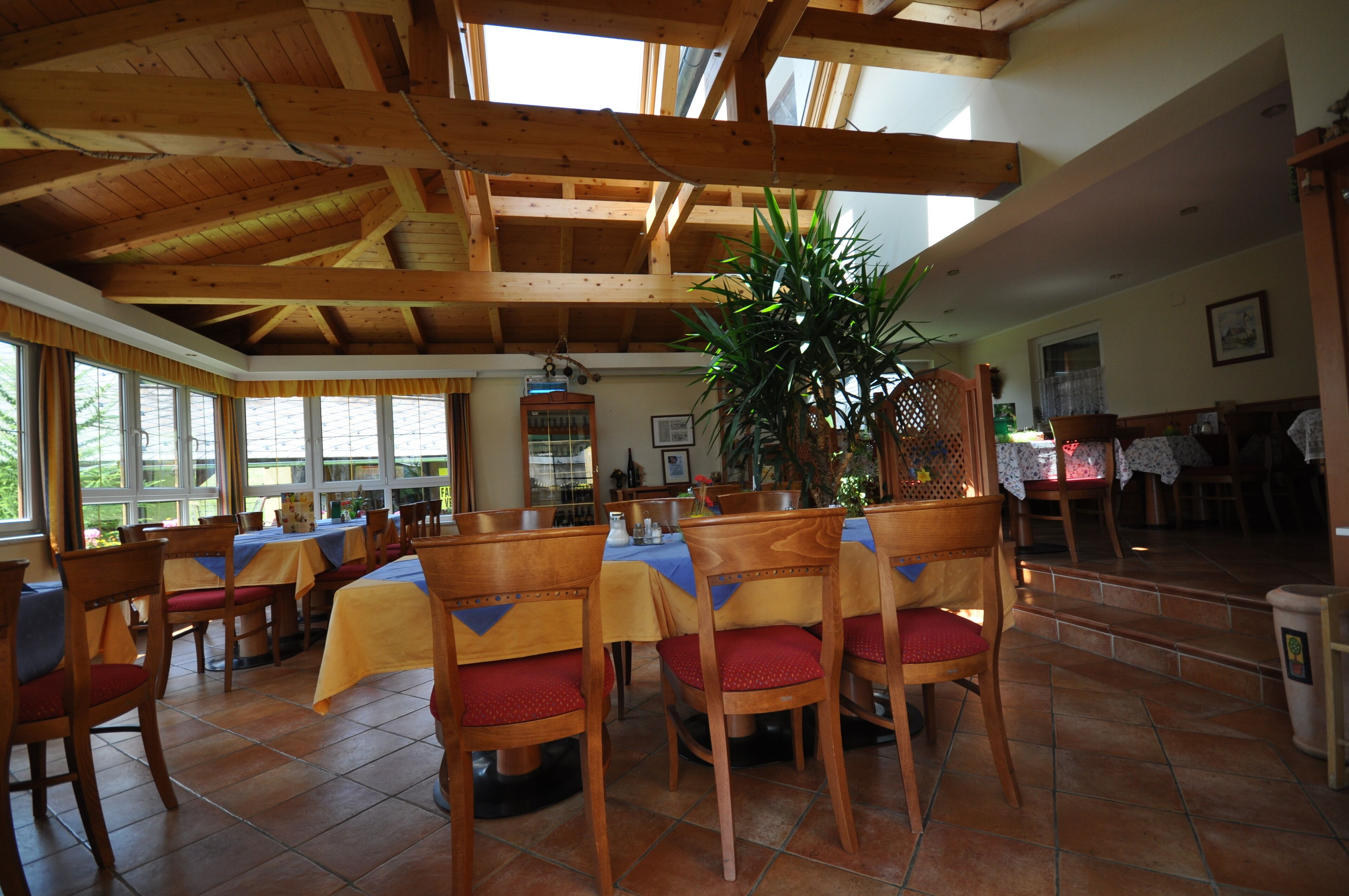 A dining area with wooden tables and chairs, large windows, and a high ceiling with exposed beams.