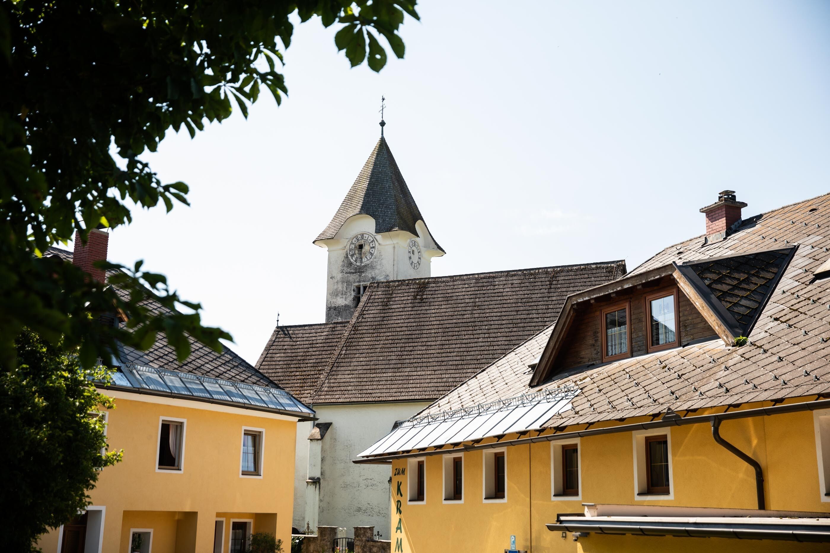 Un pueblo europeo tradicional con una iglesia que tiene una torre con reloj, edificios amarillos con techos a dos aguas y un cielo despejado.