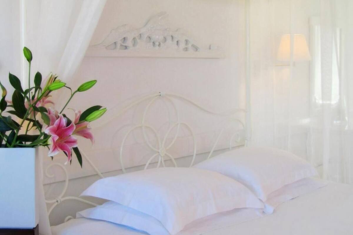 Una cama bien hecha con sábanas blancas y un florero con flores rosadas sobre una mesita de noche.