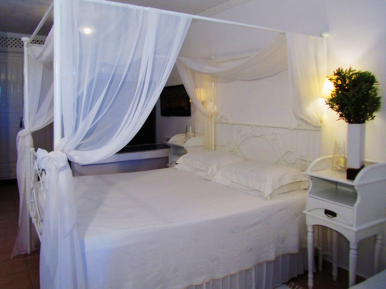 A four-poster bed with white bedding and sheer drapes, a bedside table with a lamp, and a small potted plant.