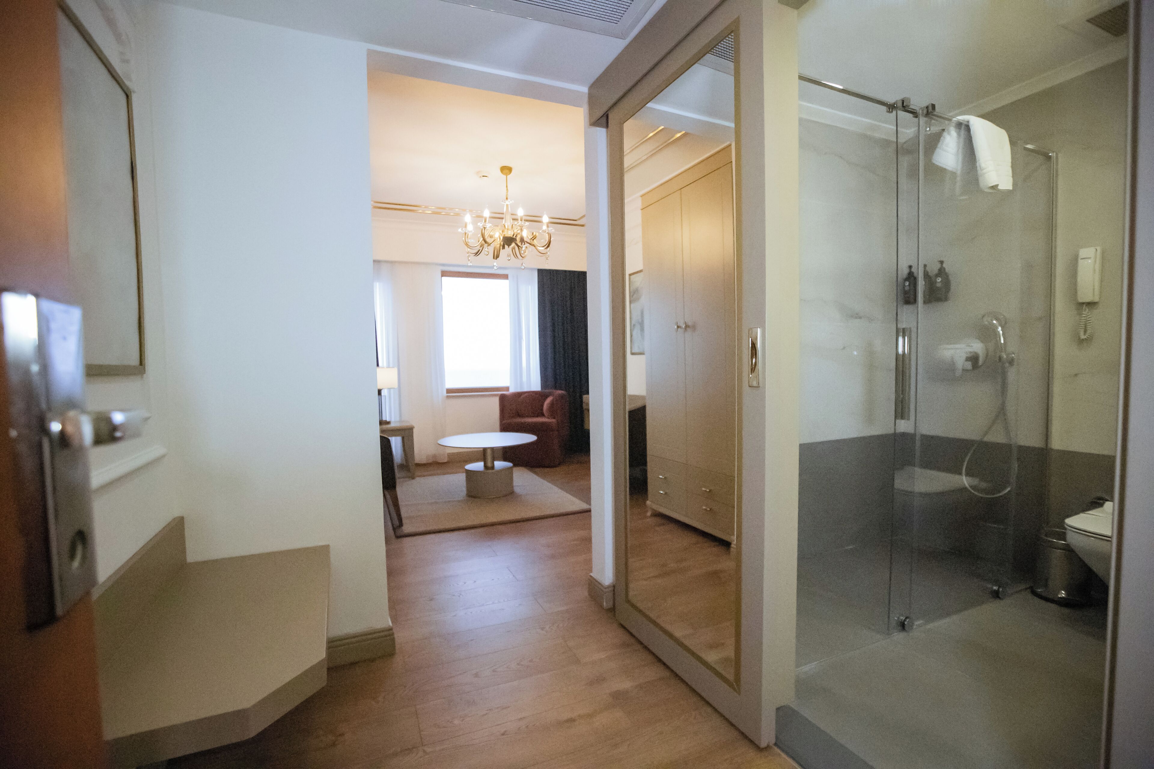 A modern hotel room with a glass shower enclosure, a wooden floor, a round table, and a chandelier.