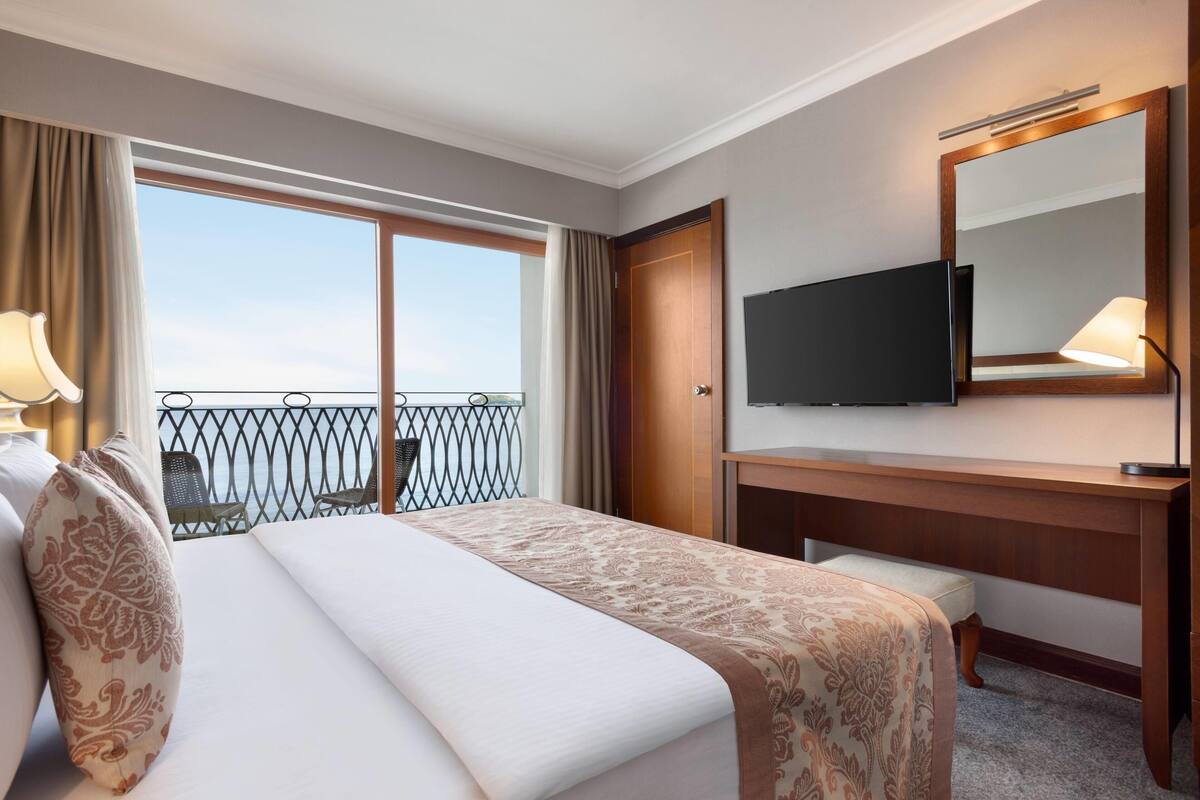 Ein Hotelzimmer mit Bett, Schreibtisch, Fernseher, einem Balkon mit Aussicht und einem Vorhangfenster.