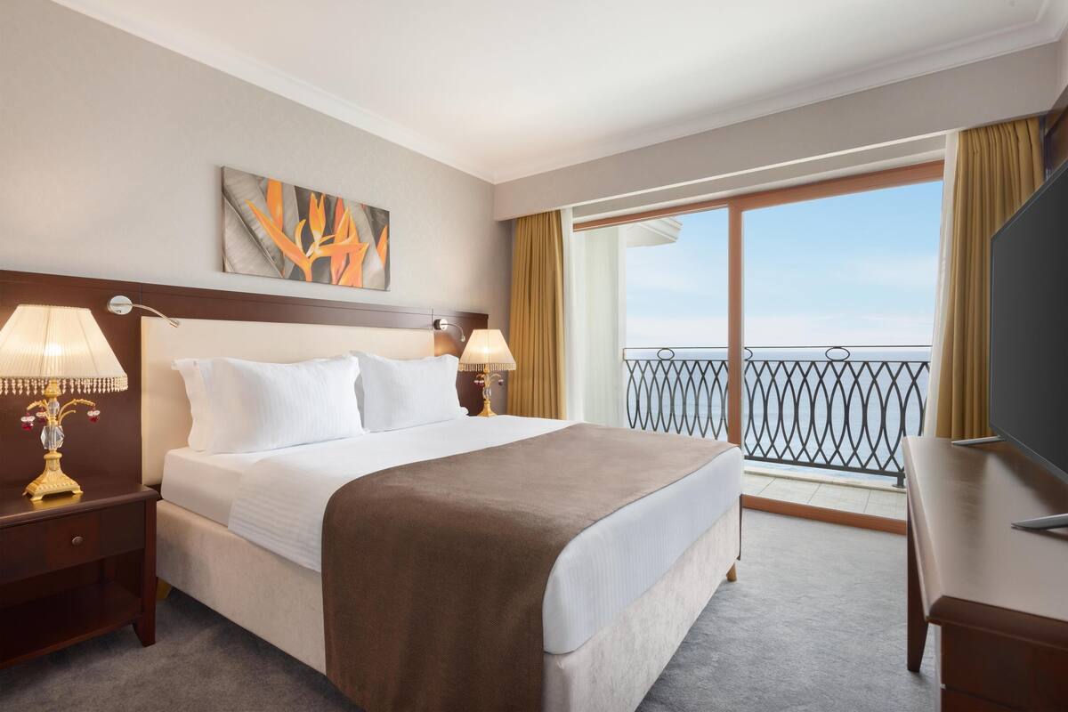 Ein Hotelzimmer mit einem großen Bett, zwei Nachttischlampen, einem Flachbildfernseher und einem Balkon mit Geländer.