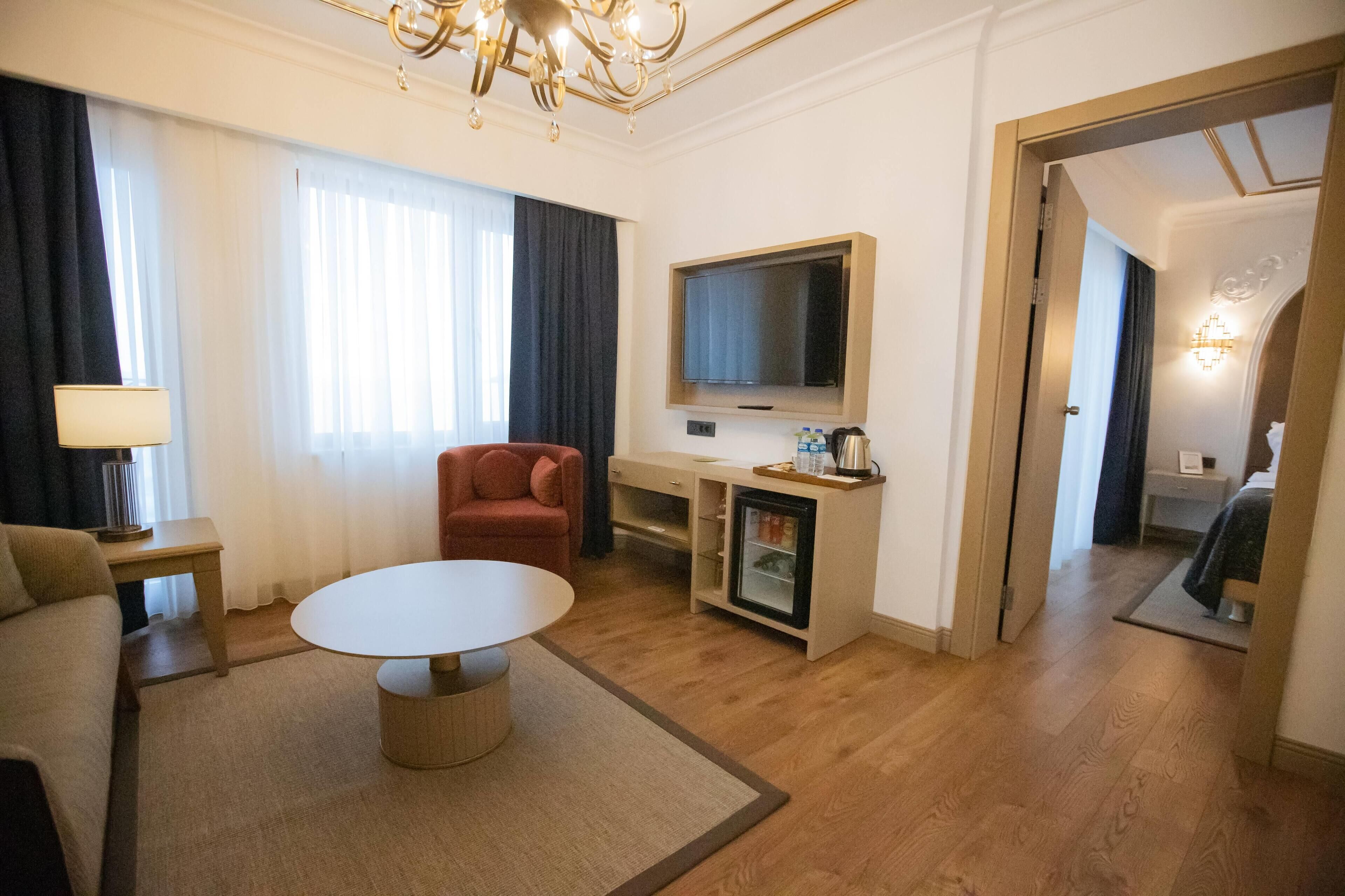 A hotel room with a round table, a red armchair, a TV, a minibar, and a bed in the adjacent room.