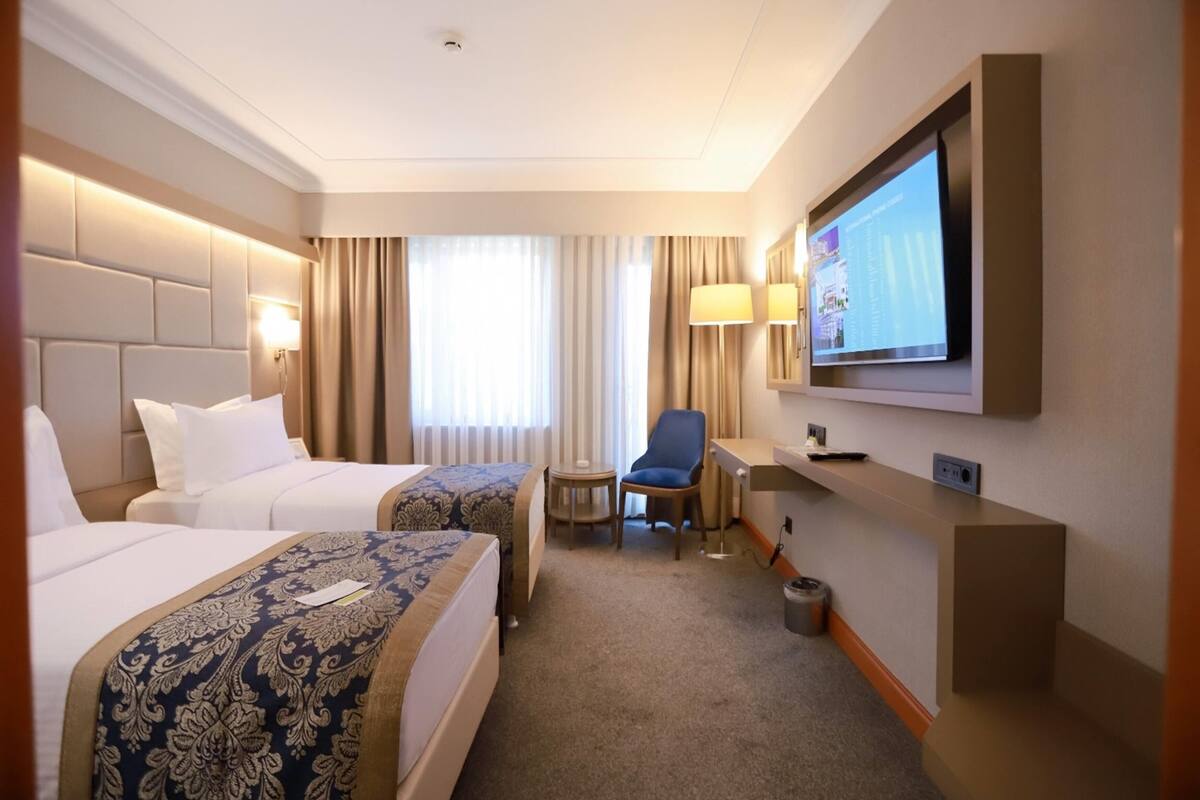 Ein Hotelzimmer mit zwei Betten, einem Schreibtisch, einem Sessel, einem Fernseher und einem Fenster mit Vorhängen.